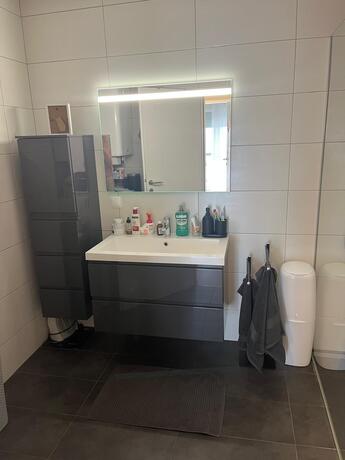 Modernes Badezimmer mit grauem Waschtischunterschrank, beleuchtetem Spiegelschrank und dunklen Fliesen